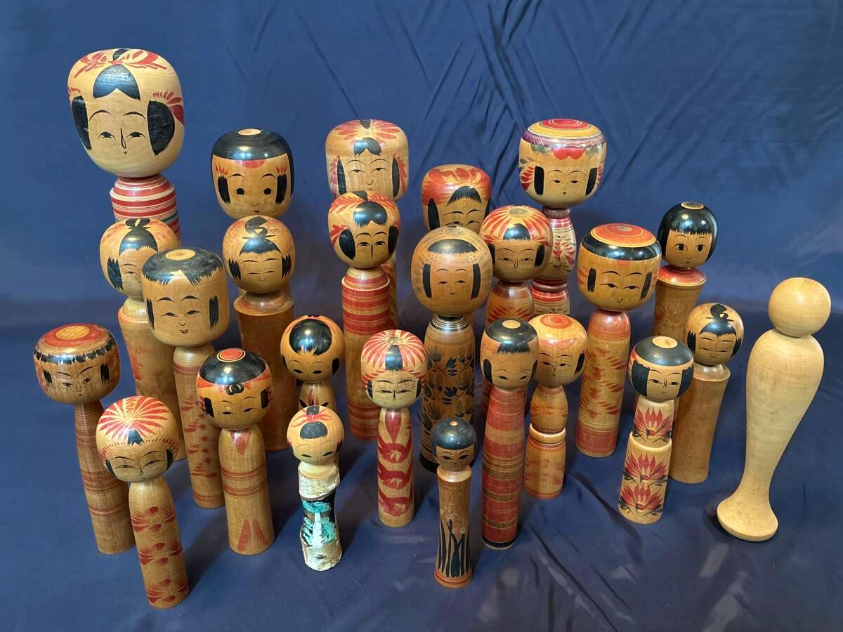 Twenty-five wooden kokeshi dolls of various sizes and painted faces arranged on a blue cloth background.