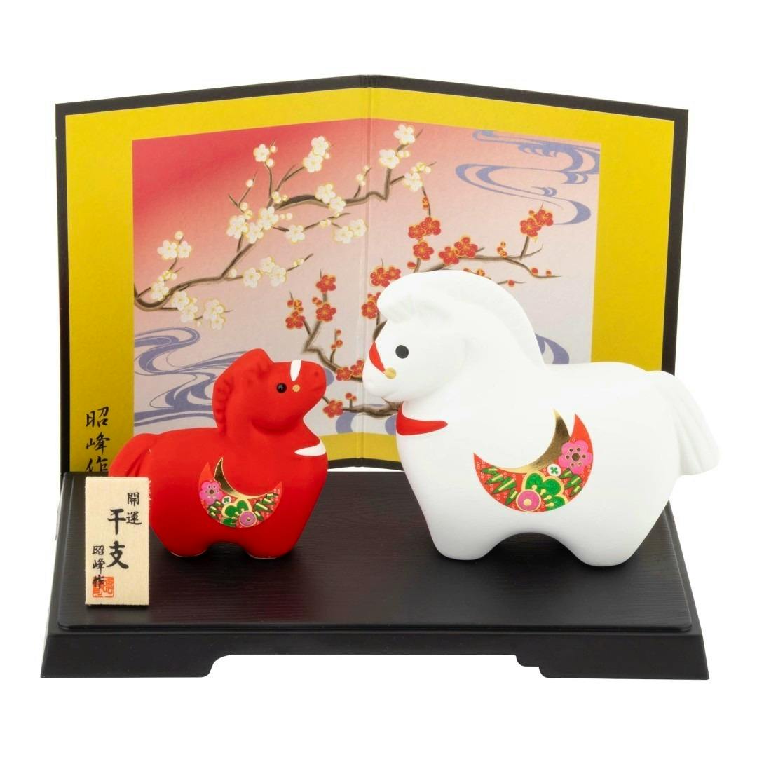 Two ceramic horse figurines (one white, one red) displayed on a black base with a decorative gold folding screen and a small wooden plaque.