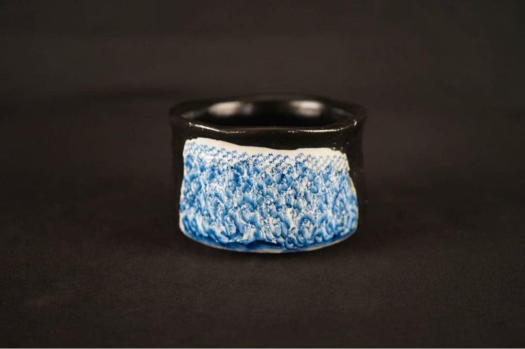 Front view of the tea bowl showing blue underglaze patterned decoration on the lower body and a black-glazed rim against a black background.