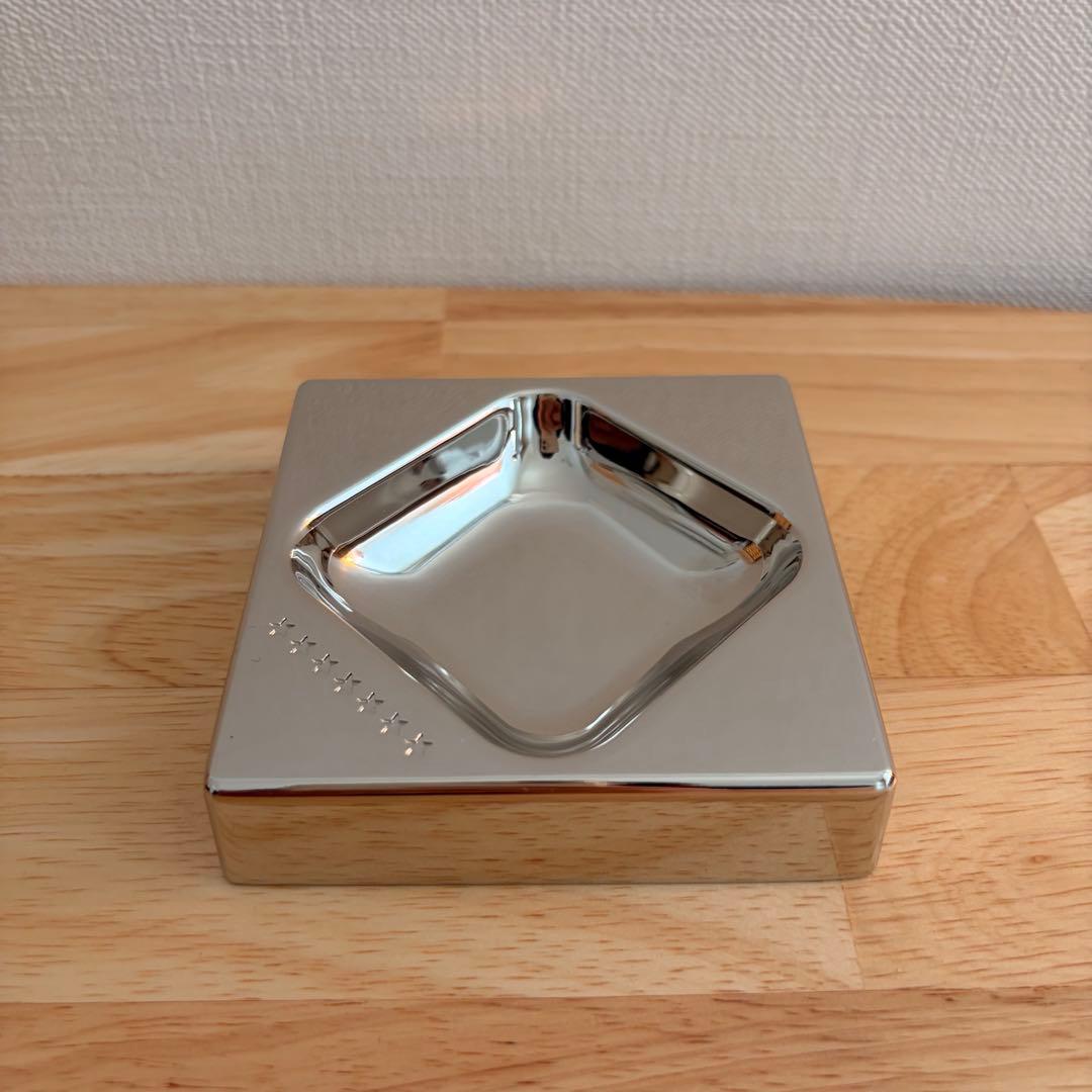 Stainless steel square tabletop ashtray with a recessed central dish and small embossed star marks, shown on a wooden surface.