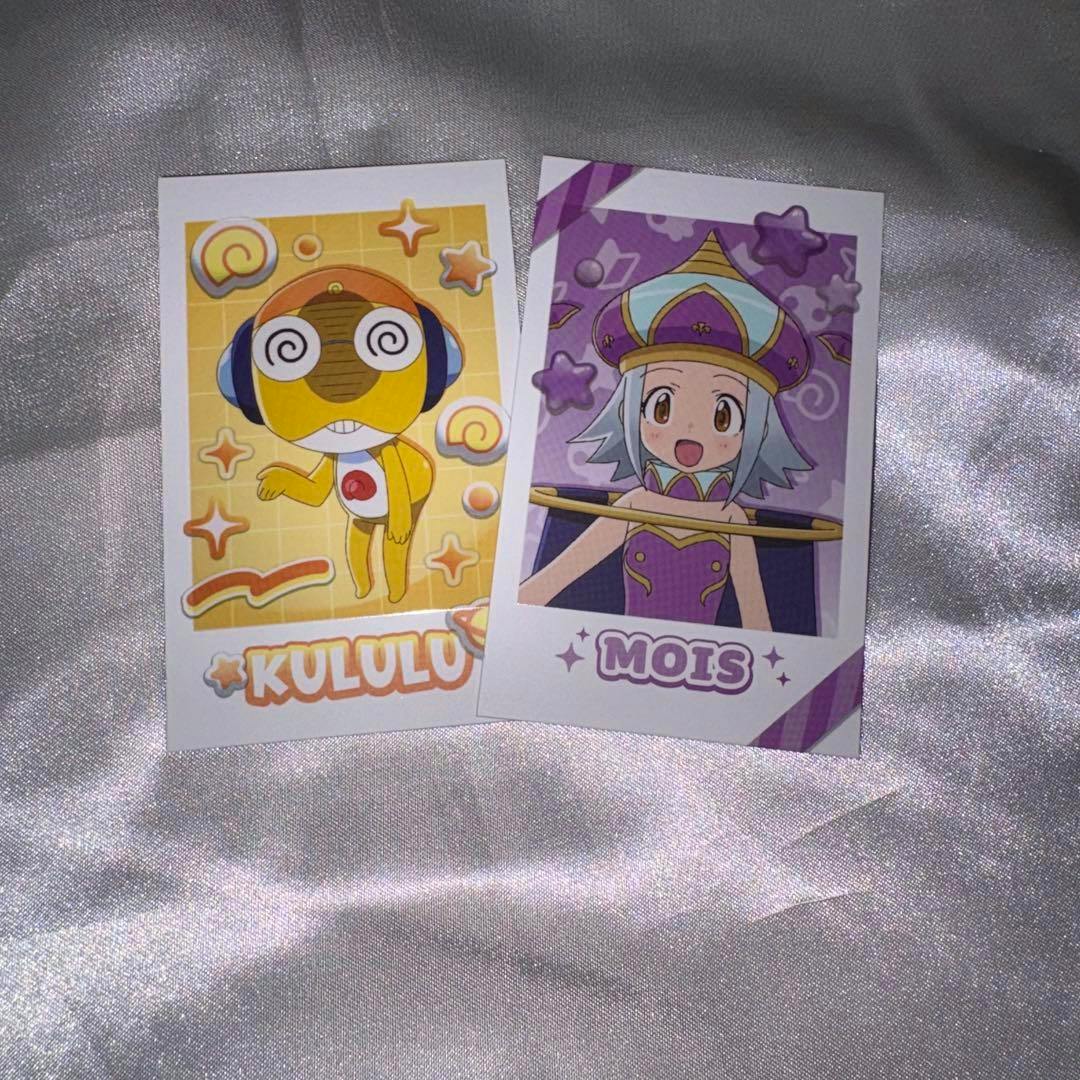 Two printed photocards showing the characters Kululu (left) and Mois (right) placed on a silver fabric background.