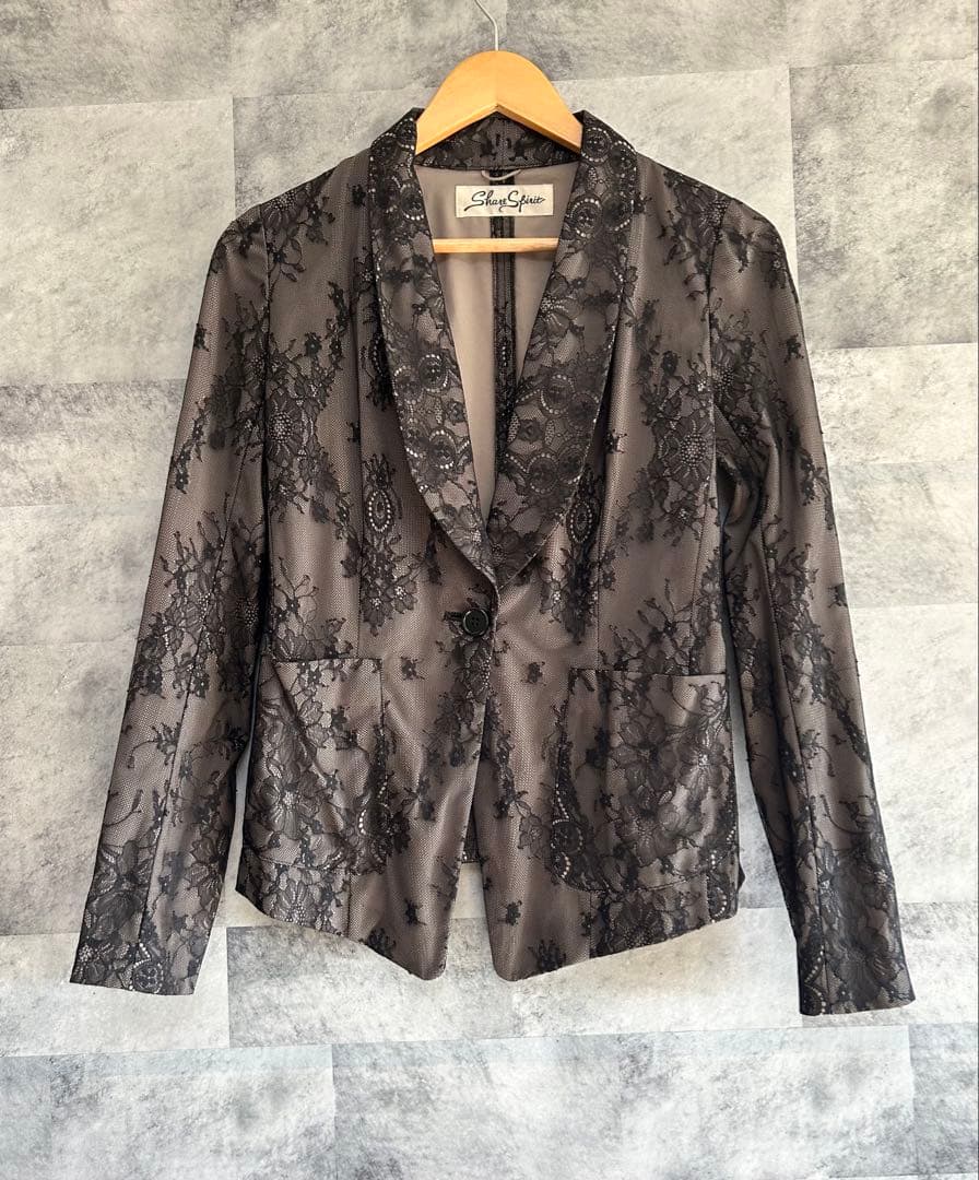 A women's polyester jacket with an all-over floral lace pattern hanging on a wooden hanger against a tiled wall.