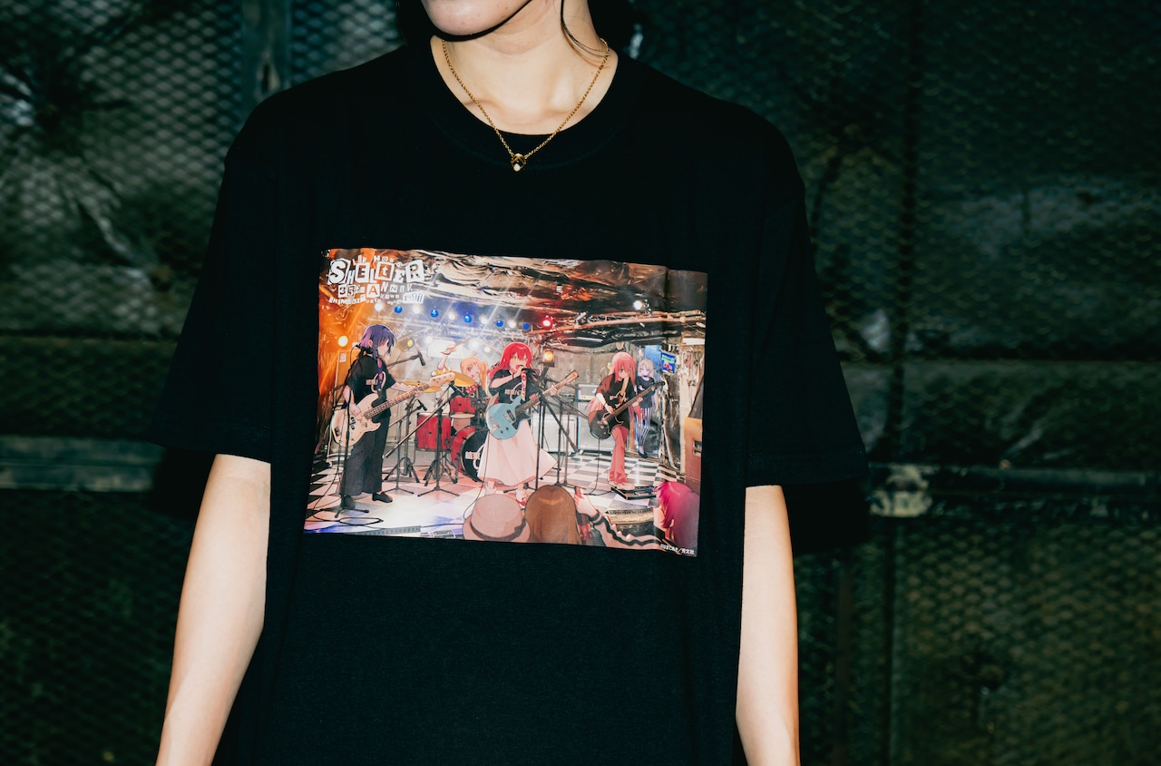 Person wearing a black cotton T-shirt showing a full-color printed illustration of a band scene on the chest.