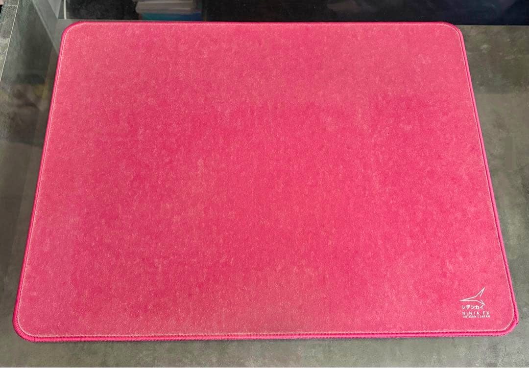 A large strawberry-pink rectangular polyester mouse pad with stitched edges and a small logo in the lower-right corner, shown on a dark surface.
