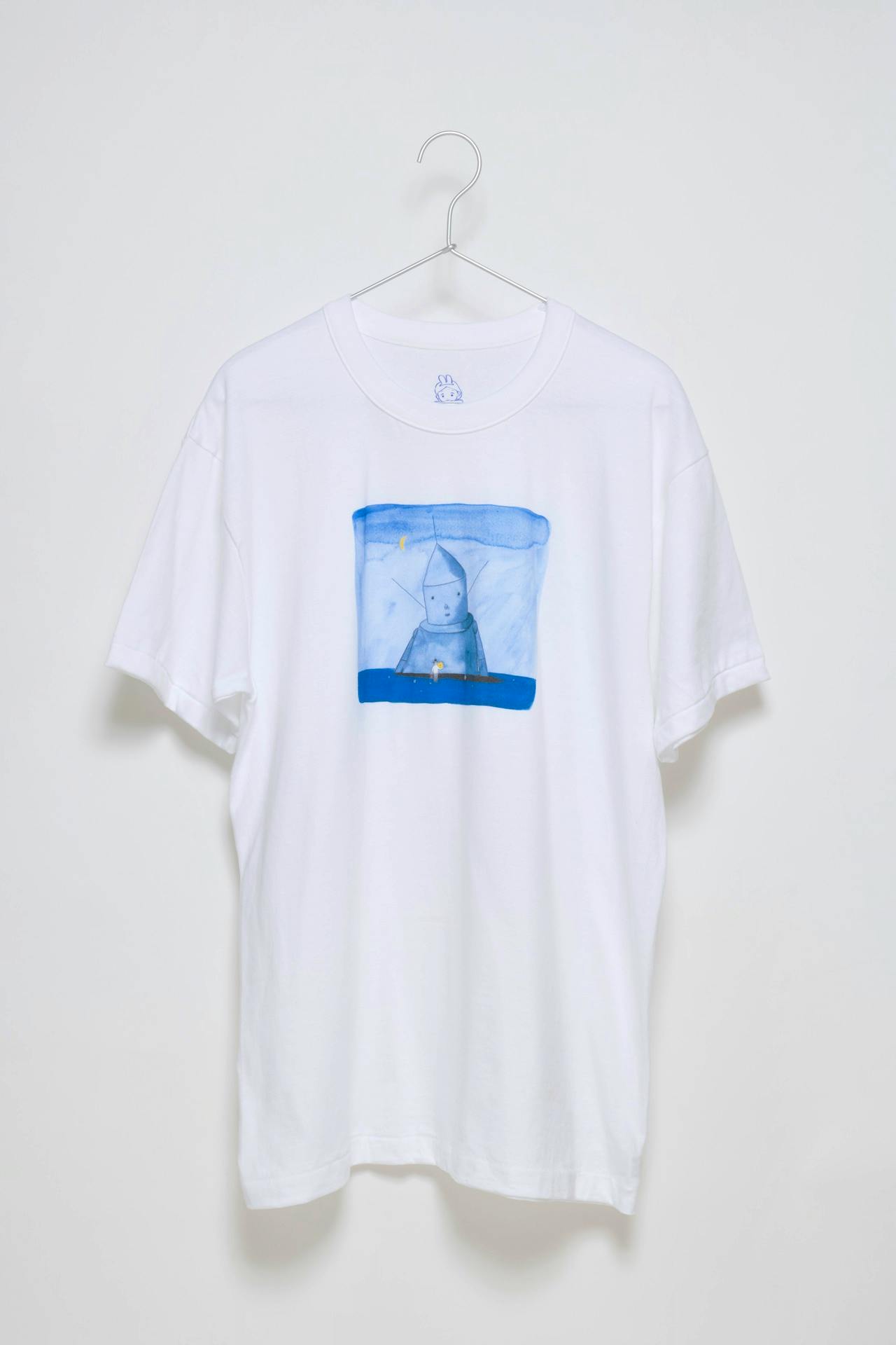 White cotton T-shirt with the blue "アーティファクト" artwork by たむらしげる printed on the front, shown hanging on a hanger.