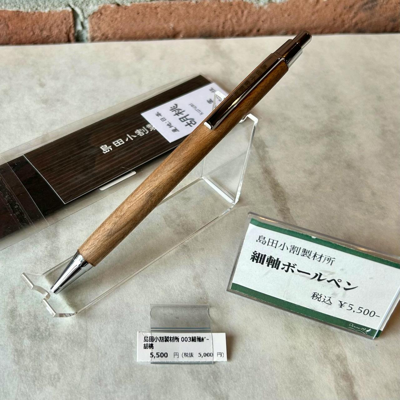 A slim walnut wood ballpoint pen with silver metal fittings displayed on a clear stand next to price tags.
