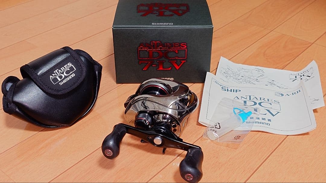 Shimano 07 Antares DC7LV right‑hand fishing reel with its box, soft case, manuals, and small parts arranged on a wooden floor.