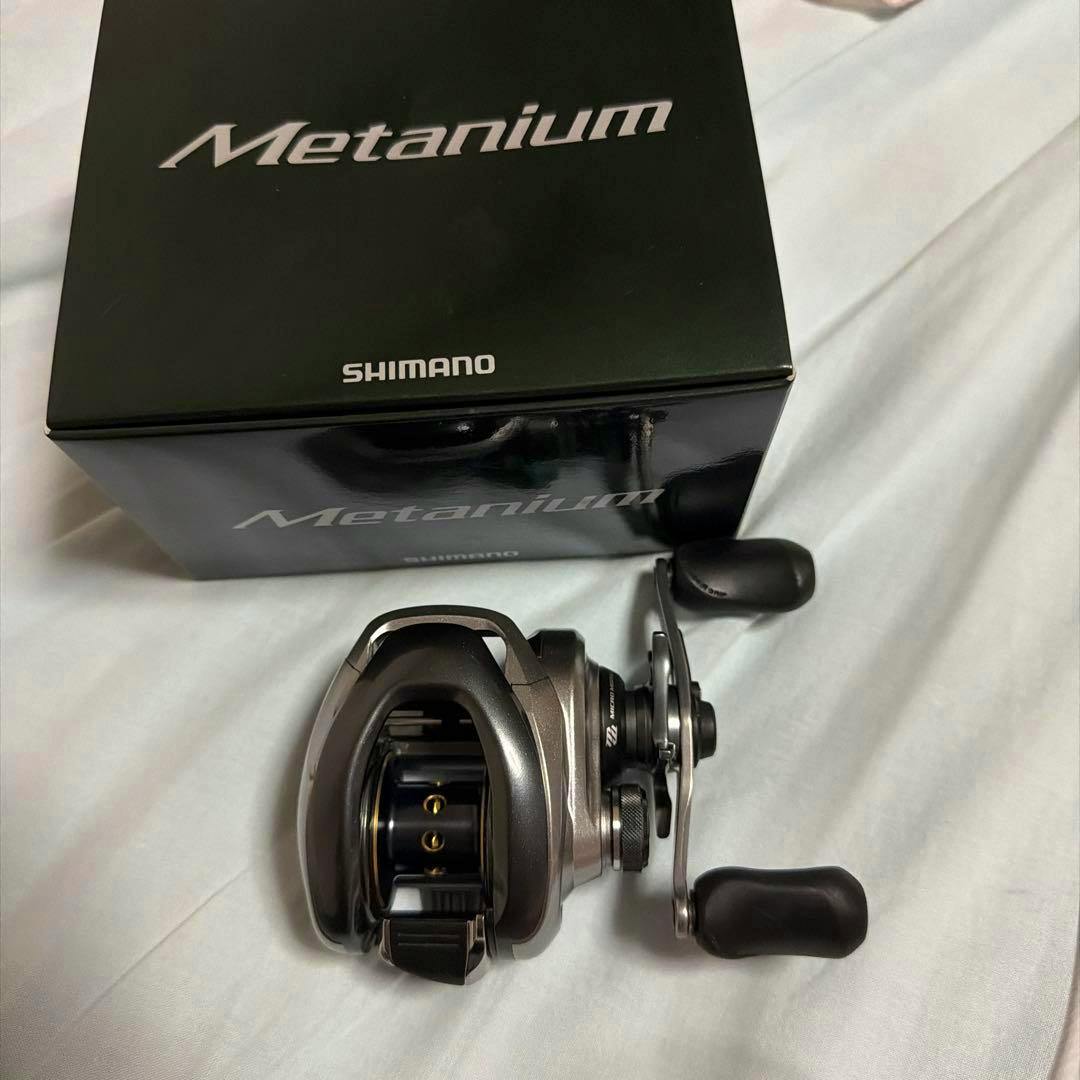 A SHIMANO Metanium baitcasting reel placed beside its Metanium box on a light fabric surface.