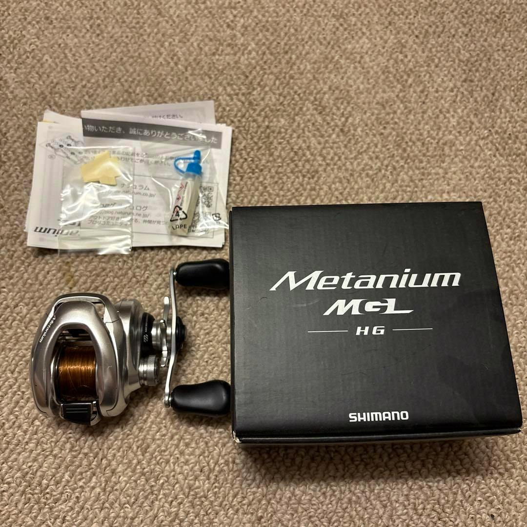A Metanium MGL HG fishing reel shown next to its box, manuals, and small accessories on a carpeted surface.