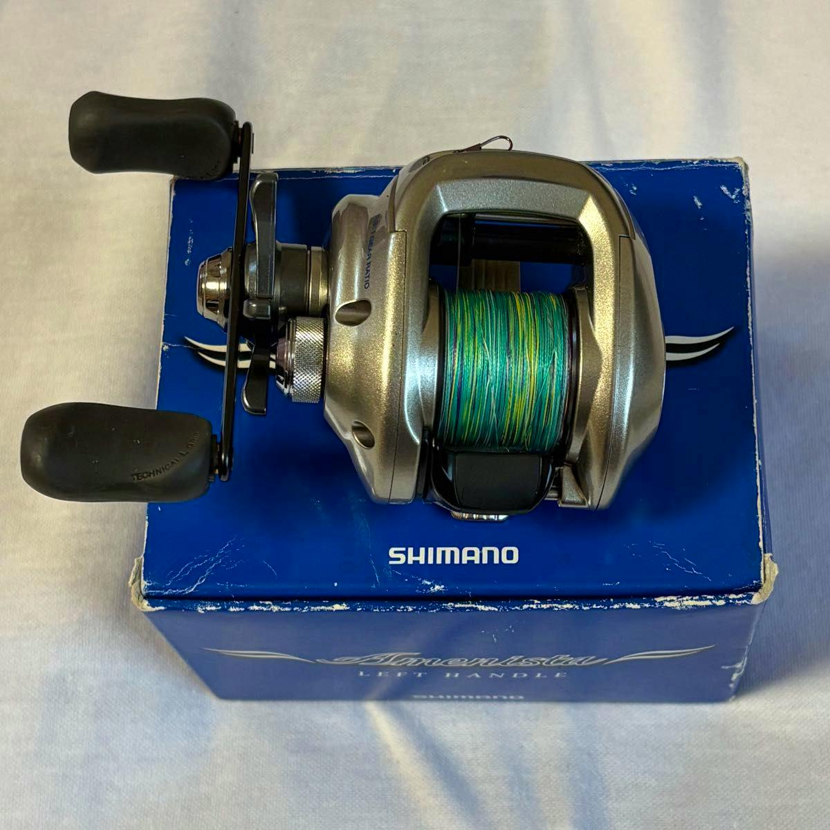 Shimano Amerista left‑handle baitcasting reel spooled with green line placed on its blue Shimano box.