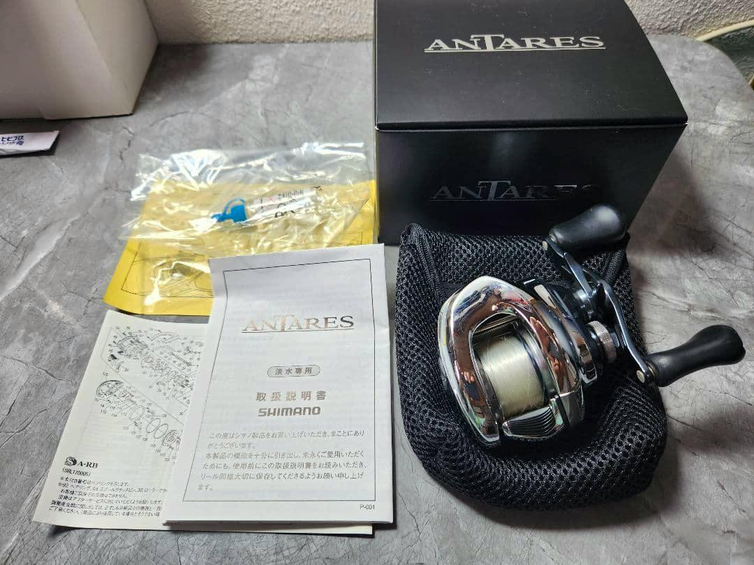 Antares baitcasting fishing reel displayed with its box, manuals, and a protective pouch on a table.