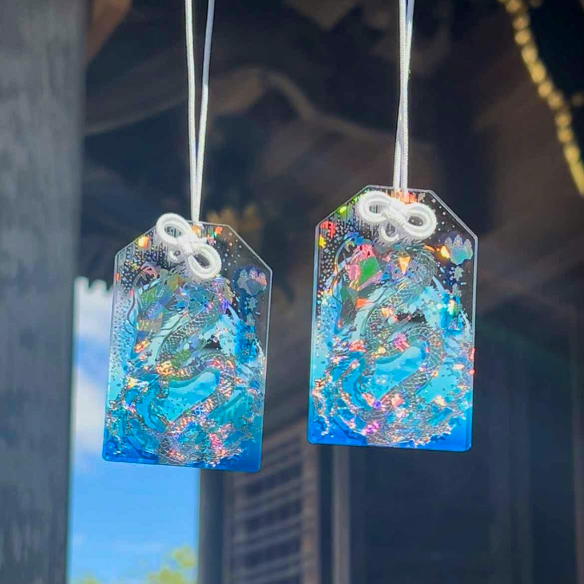 Two blue omamori amulets with holographic dragon foil hanging by white cords against a blurred background.