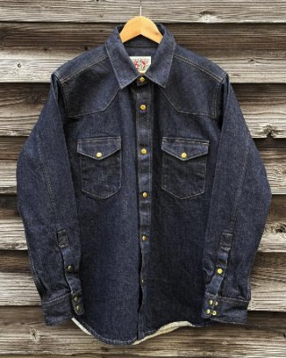 Front view of the Ship John Townes indigo denim men's shirt hanging on a wooden hanger against a wood plank background.