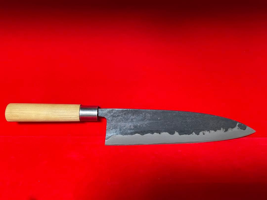 Full-length chef knife with a light wooden handle and a dark patterned blade displayed on a red background.