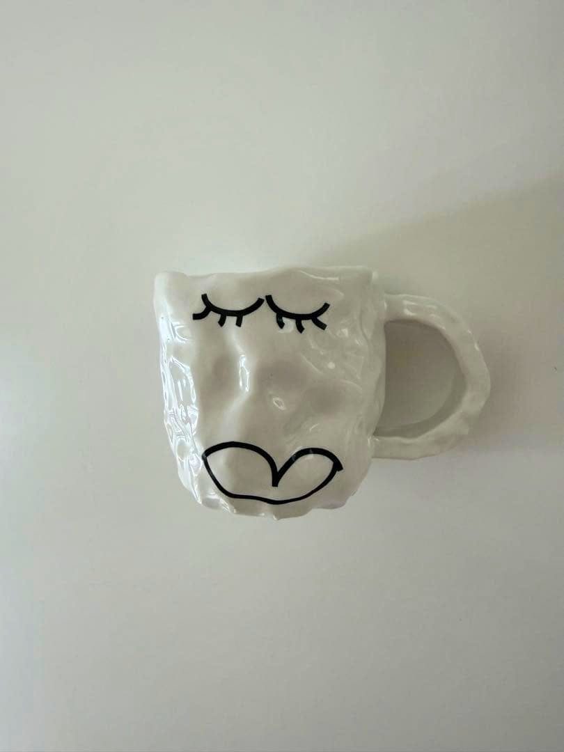 White ceramic mug (SHOKKI 平山昌尚さんコラボマグカップ) with a black eyelash and lip face design and a handle on a plain background.