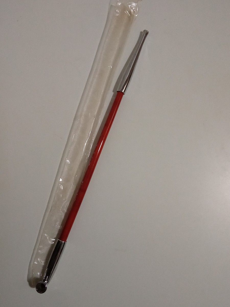 A long kiseru smoking pipe with a red lacquered stem and metal fittings lying next to a plastic sleeve on a white surface.