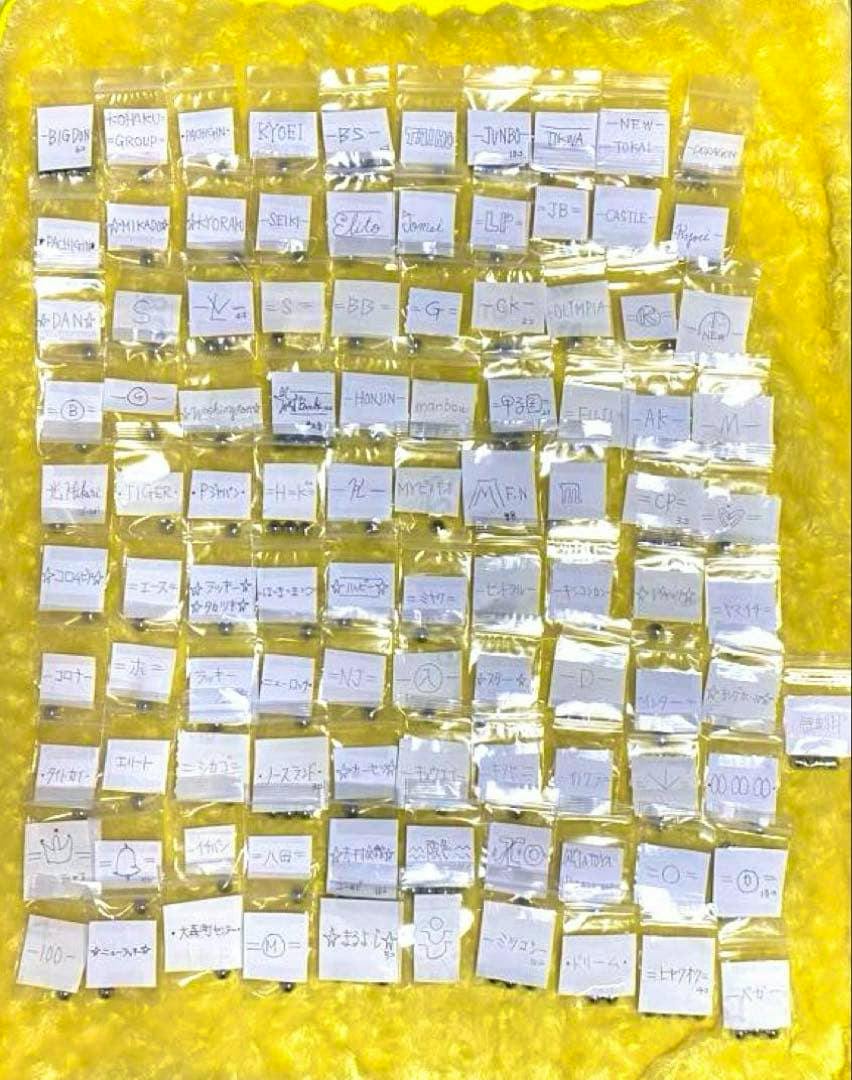 Many small sealed plastic bags each containing a metal pachinko ball with handwritten labels arranged on a yellow background.