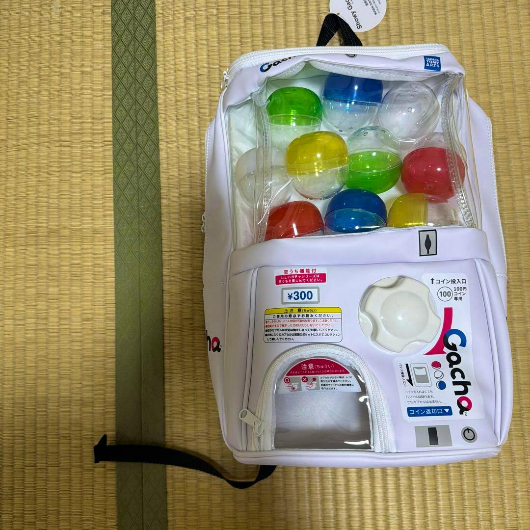 White Showy Gacha2 gacha-machine style backpack with colorful plastic capsules visible in the clear top compartment, placed on a tatami mat.