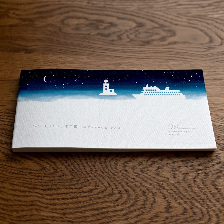 A closed silhouette message pad titled 「旅立ちの夜」 showing a night-sky design with a lighthouse and ship on a wooden surface.