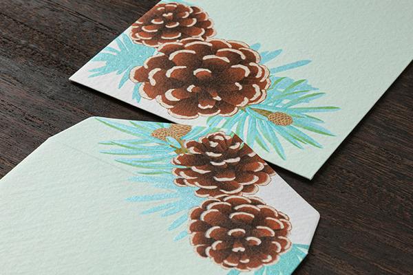 Close-up photo of a textured envelope showing a pinecone wreath design printed on pale paper.