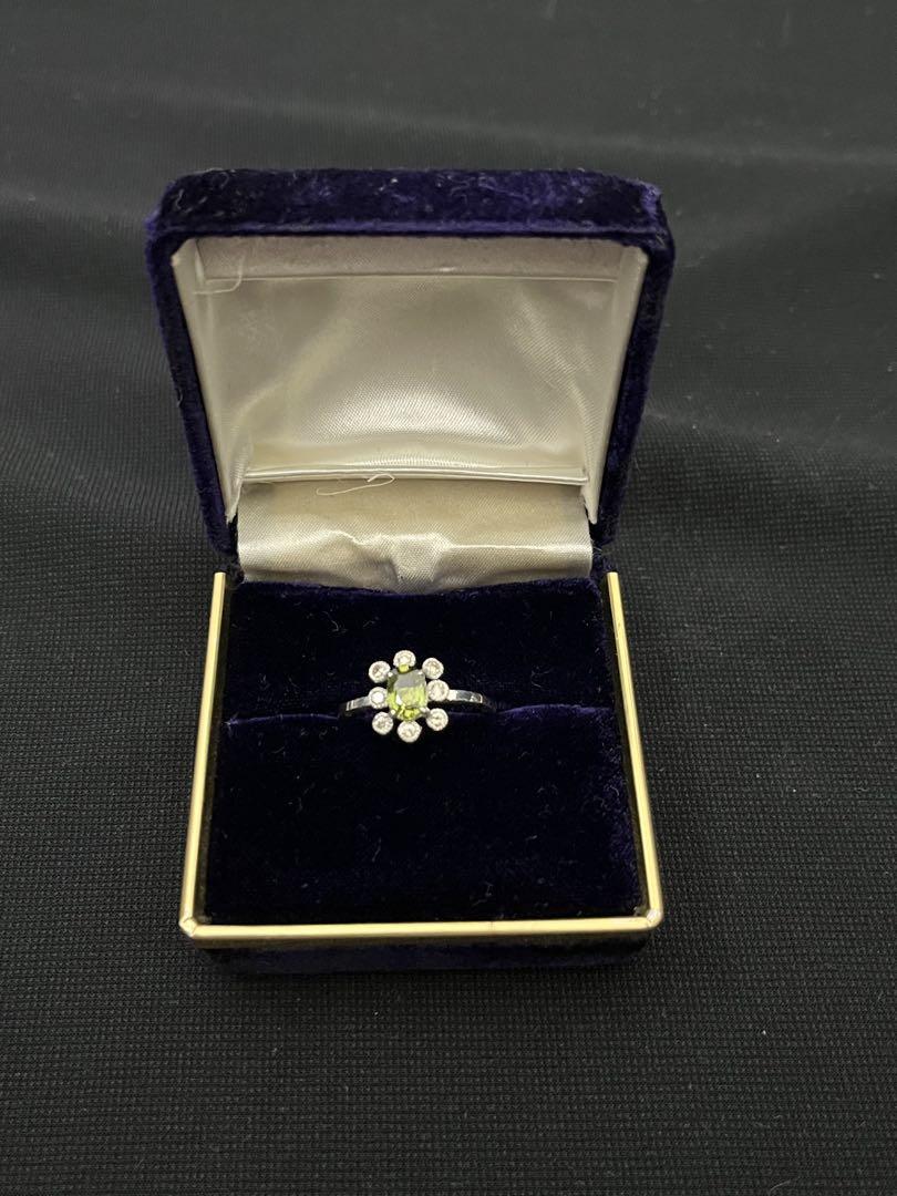Silver ring with an oval green center stone surrounded by small round clear stones, displayed in a dark velvet ring box.