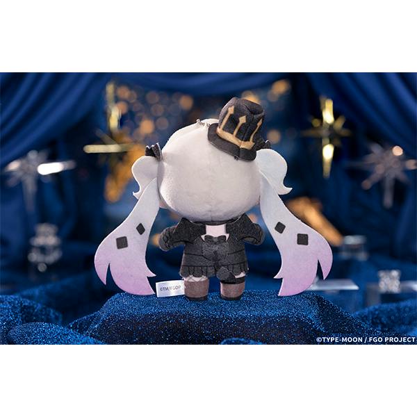 Back-view photograph of a small plush keychain charm wearing a dark outfit with pink-tinged twin tails and a small hat, displayed on a blue sparkly surface.