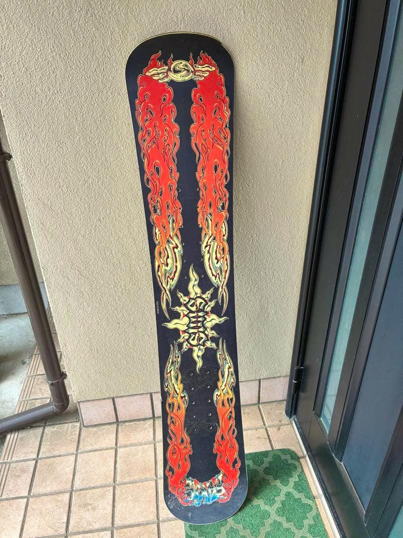 A SIMS snowboard showing the top sheet with red-orange flame graphics leaning against a wall on a tiled floor.