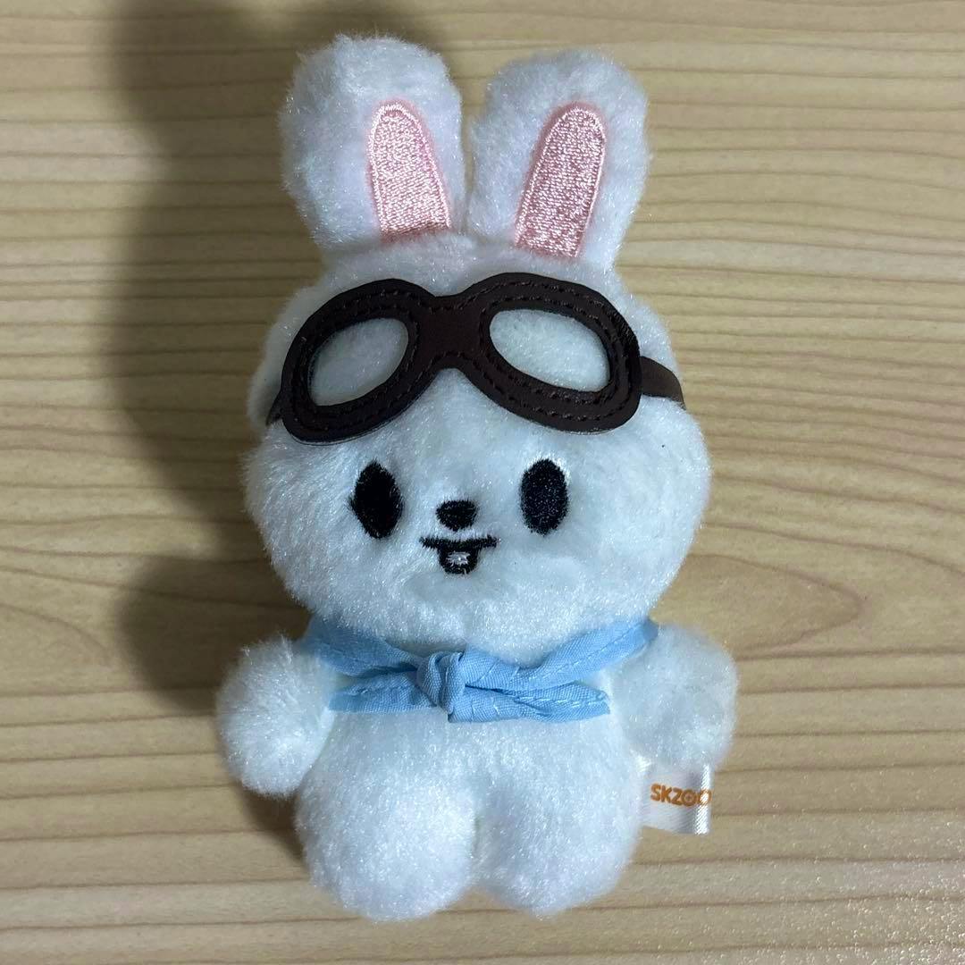 Front view of a white SKZOO Leebit pilot plush rabbit wearing brown goggles and a blue neckerchief on a wooden surface.