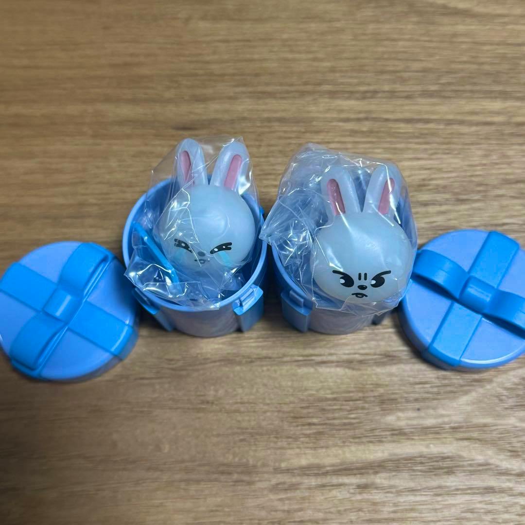 Two SKZOO Leebit rabbit plastic toys sealed in clear bags and placed inside blue round plastic containers on a wooden surface.