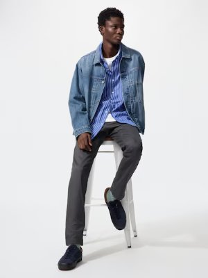 Male model seated on a white stool wearing gray slim-fit chino pants and a denim jacket.