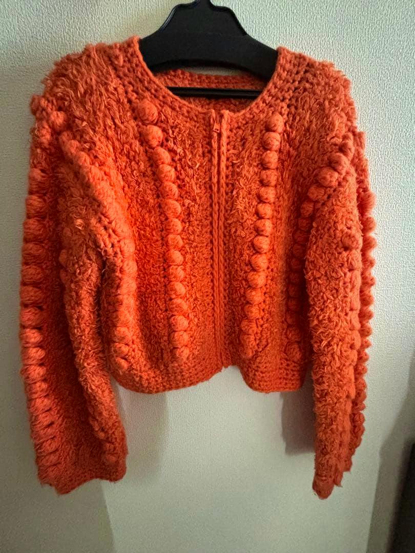 Orange fluffy blouson with vertical pom‑pom knit details and a front zipper hanging on a black hanger against a wall.