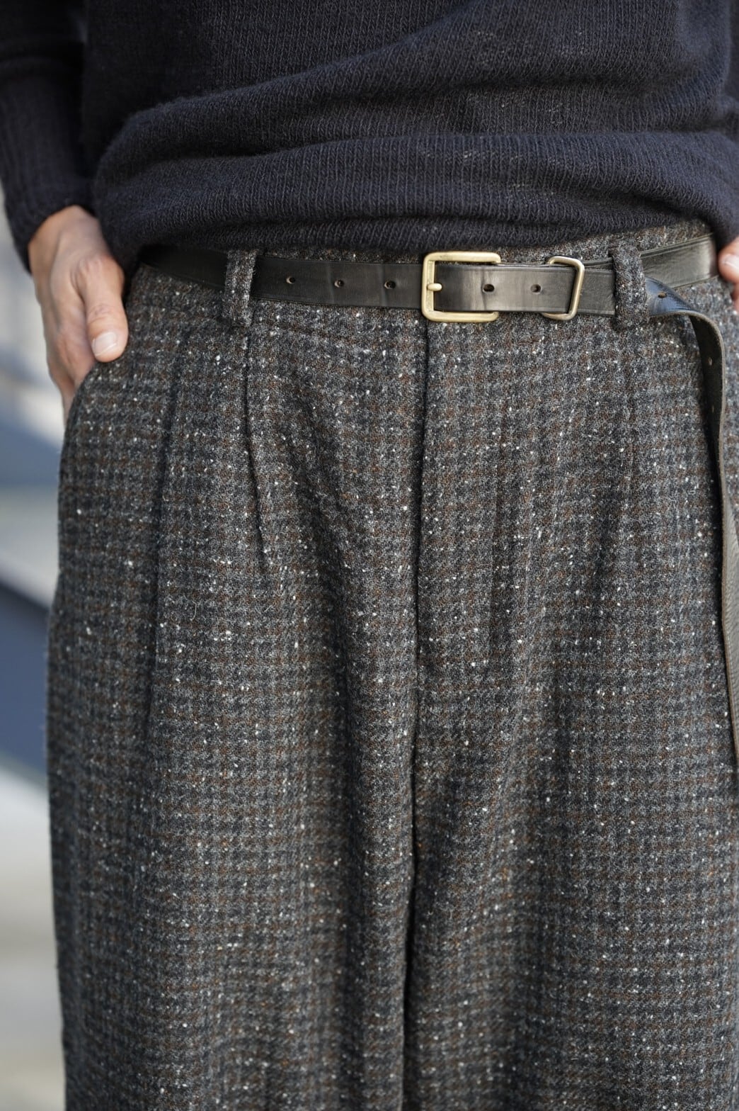 Black cow leather belt with a metal buckle worn around the waist over gray checked trousers.