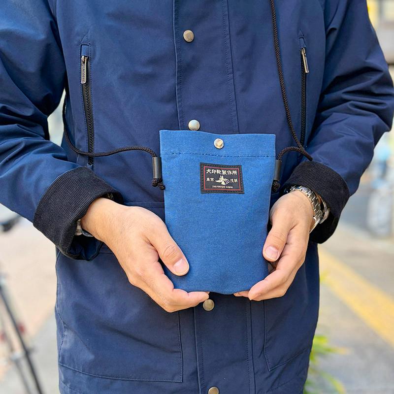 A person wearing and holding a blue cotton canvas smartphone pouch (スマホポシェット) on a cord in front of a navy jacket.