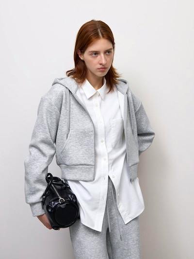 Model wearing a light gray full-zip hooded sweatshirt over a white button-down shirt and matching gray sweatpants, holding a black bag.