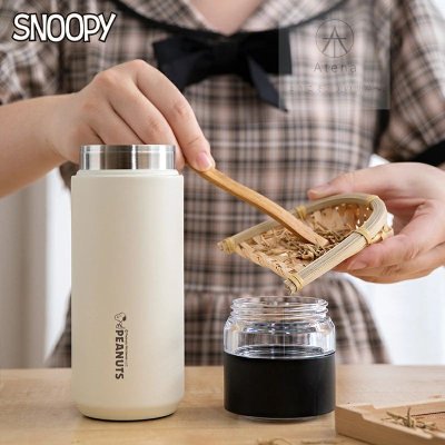 Beige Snoopy stainless steel tumbler on a table with a person holding a tea strainer next to a jar.