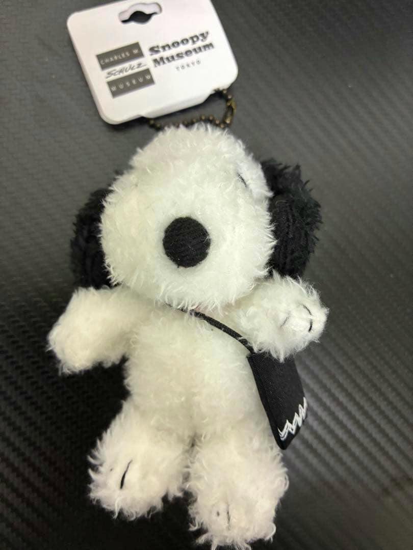 Front view of the mini Snoopy plush strap with black ears, black nose, small black bag, and attached Snoopy Museum hang tag.