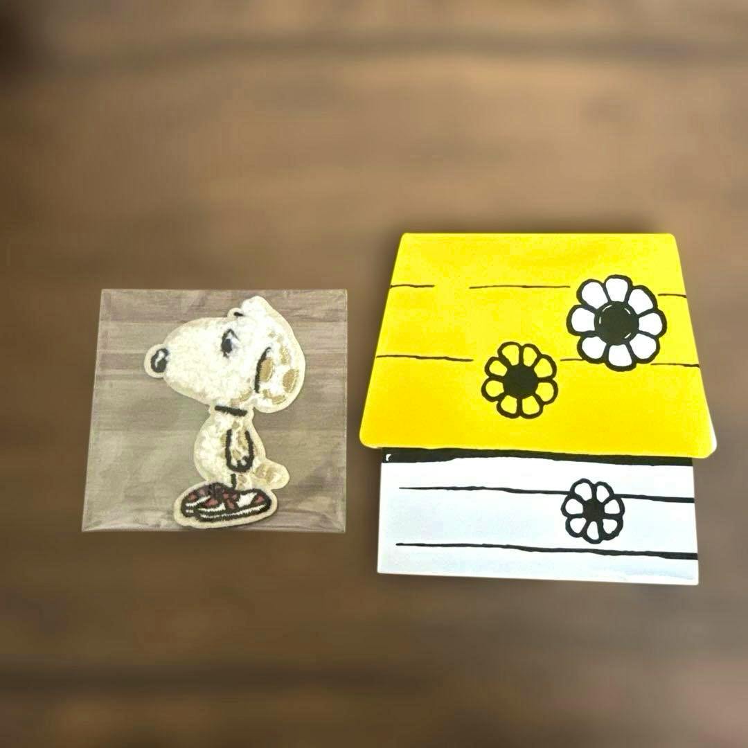 Clear plastic-wrapped Snoopy embroidered paper sticker displayed beside a yellow-and-white floral backing on a wooden surface.