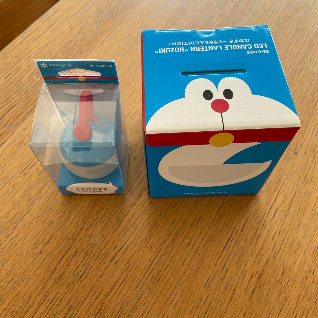 Unopened Doraemon-themed Snow Peak LED table lamp in clear plastic packaging next to its Doraemon box on a wooden table.