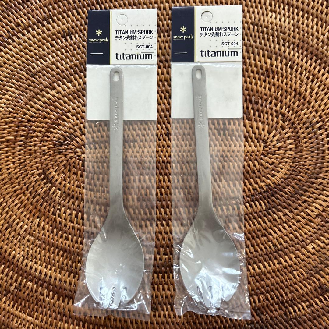 Two Snow Peak titanium split-tip spoons (SCT-004) in clear retail packaging shown side by side on a woven mat.