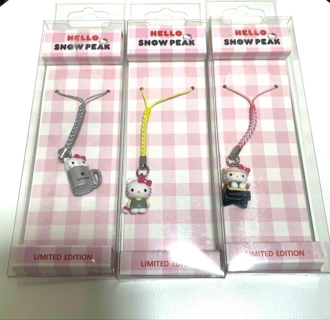 Three Hello Kitty × Snow Peak keychain strap figures shown in clear boxed packaging on a pink checkered backing.