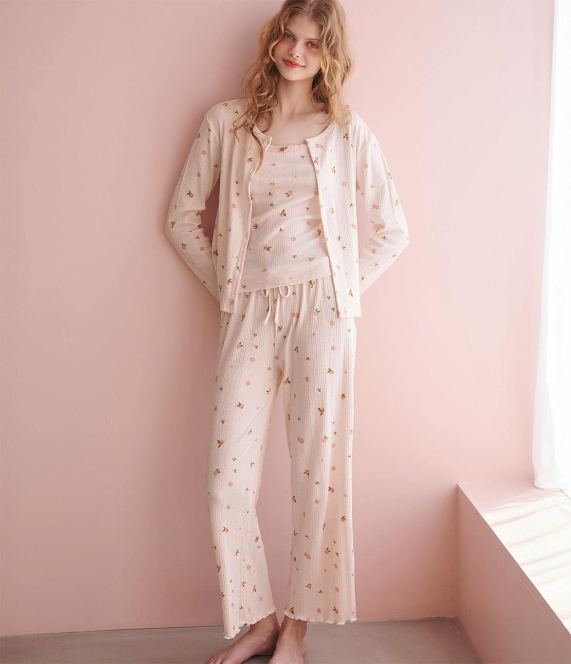 Model wearing the three-piece roomwear set (tank top, buttoned cardigan, and drawstring pants) in a light floral ribbed fabric against a pink wall.