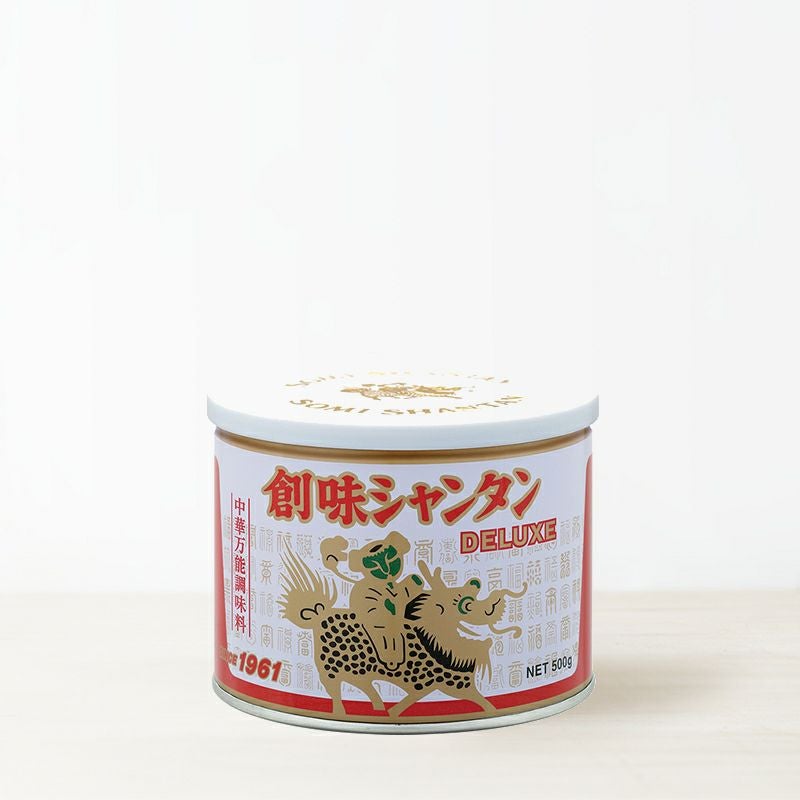 A 500g can of 創味シャンタンDX (Deluxe) with a white lid and a red-and-white label showing a stylized dragon illustration.
