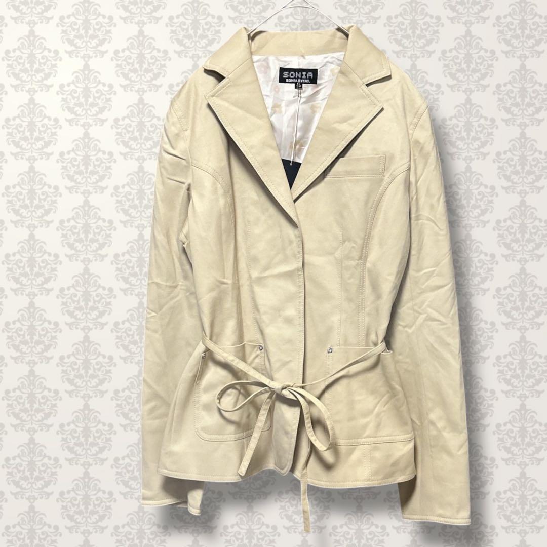 Beige Sonia Rykiel short-length women's trench coat (rayon) hanging on a hanger against a patterned wall.
