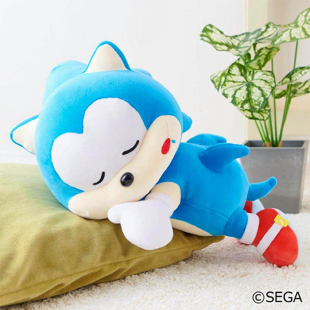 A blue Sonic plush toy lying on its side with closed eyes on a green cushion, with a potted plant in the background.