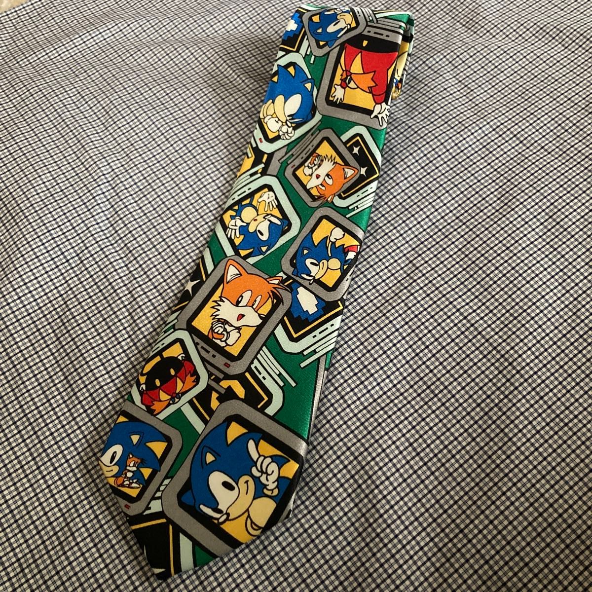 A polyester necktie laid flat on checkered fabric, with an all-over green pattern showing Sonic the Hedgehog characters in small square frames.