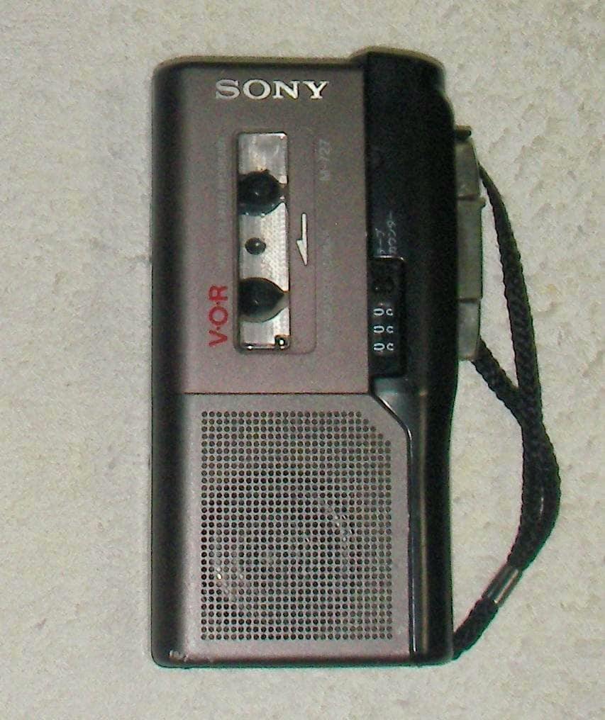 Front view of the Sony M-727 microcassette recorder showing the cassette window, speaker grille, controls, and a wrist strap.