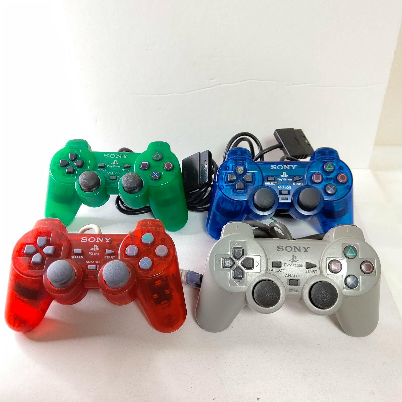Four PlayStation (PS/PS2) DualShock controllers in green, blue, red, and silver arranged on a white background.
