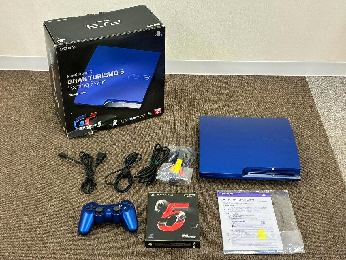 Blue Sony PlayStation 3 console with a matching blue controller, cables, Gran Turismo 5 game, manuals, and the original box arranged on a floor.