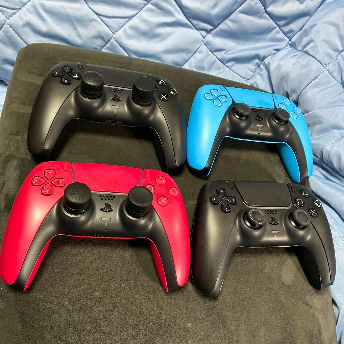 Four PS5 DualSense controllers (black, blue, red, and black) arranged on a fabric surface showing the front-facing controls.