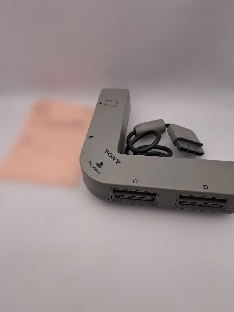 Grey Sony PlayStation PS1 multitap adapter (SCPH-1070) showing the L-shaped unit with controller ports and attached cable on a white background.